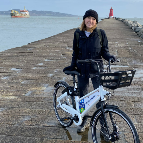A woman stands in front of a red lighthouse holding a bicycle.